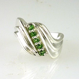 May include: A silver-toned ring featuring a central diagonal channel set with five small, round, vibrant green gemstones. The band has a polished, smooth, and undulating wave-like design that curves around the finger, highlighting the contrast between the metallic surface and the green stones.