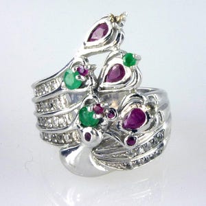 May include: A silver ring featuring a unique design with ruby and emerald gemstones. The ring has a layered appearance, with multiple bands adorned with small diamonds. The design incorporates stylized leaf or bird-like shapes, with the gemstones set within.