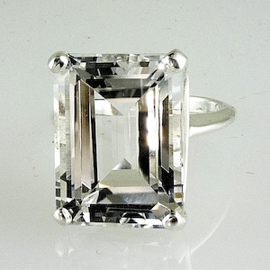 Könnte beinhalten: Ein Ring mit einem klaren, rechteckig geschliffenen Edelstein und einem silbernen Band. Der Edelstein ist mit vier kleinen silbernen Zinken an jeder Ecke gefasst. Das Design des Rings zeigt einen großen, klaren und facettierten Stein, der Licht reflektiert.
