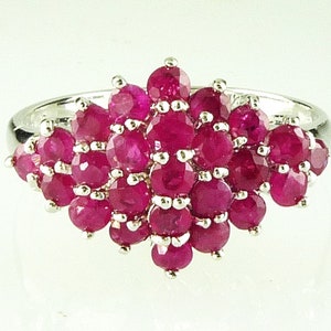 May include: A cluster ring featuring numerous round, deep red gemstones set in a silver band. The gemstones are arranged in a dense, layered pattern, creating a sparkling, eye-catching design. The ring is set against a plain white background.