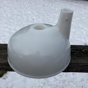 Puede incluir: Un embudo de cerámica blanca con base redondeada y boquilla. El objeto tiene un acabado liso y brillante y un pequeño agujero en la parte superior de la base. La boquilla se extiende hacia arriba y hacia un lado. El embudo está sobre una superficie de madera.