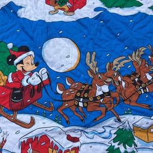 Puede incluir: Una tela acolchada con temática navideña con Mickey Mouse con un traje de Papá Noel conduciendo un trineo tirado por renos. Pluto está en el trineo. El fondo es azul con una luna llena, nieve y casas de dibujos animados.