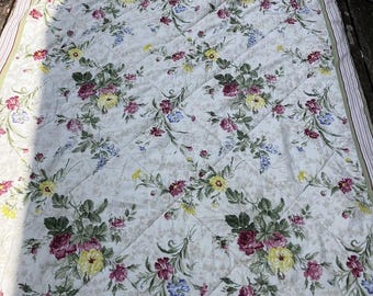 Könnte beinhalten: Eine rechteckige Steppdecke mit cremefarbenem Hintergrund und einem wiederkehrenden Blumenmuster aus roten, gelben und blauen Blüten mit grünen Blättern. Der Stoff hat eine rautenförmig gesteppte Textur und einen gestreiften Rand in gedeckten Tönen.