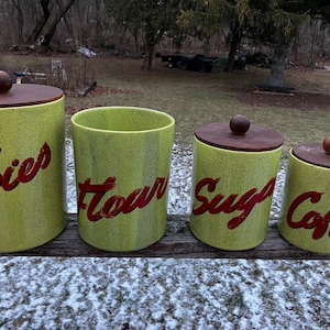 Puede incluir: Un juego de cuatro botes de cocina vintage. Los botes son de color verde claro con letras rojas que dicen "Cookies", "Flour", "Sugar" y "Coffee". Cada bote tiene una tapa marrón oscuro con un pomo de madera.