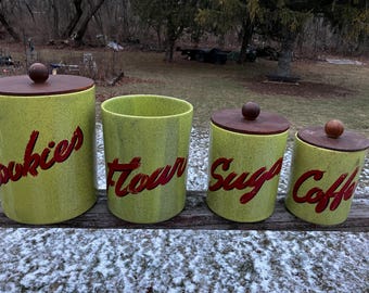 Könnte beinhalten: Ein Set aus vier Vintage-Küchenbehältern. Die Behälter sind hellgrün mit roter Schrift, die "Cookies", "Flour", "Sugar" und "Coffee" lautet. Jeder Behälter hat einen dunkelbraunen Deckel mit einem Holzknauf.