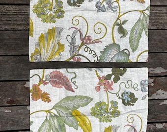 Könnte beinhalten: Zwei rechteckige Tischsets aus Stoff mit einem botanischen Druck auf weißem Hintergrund. Das Muster zeigt verschiedene grüne Blätter, gelbes Laub und Gruppen kleiner rosa, blauer und grauer Blüten, die durch dünne, gewundene braune Ranken verbunden sind.