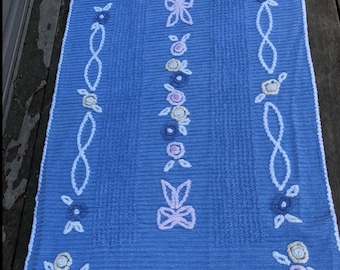 Könnte beinhalten: Eine rechteckige blaue Badematte mit einer strukturierten, gerippten Oberfläche und einem weißen Häkelrand. Die Matte ist mit gestickten Blumenmotiven in Rosa-, Gelb- und Blautönen sowie weißen schleifenartigen Mustern und Schleifen verziert.