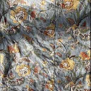 Puede incluir: Textil con estampado floral que presenta grandes flores amarillas y blancas con detalles marrones y naranjas sobre un fondo azul claro. El diseño incluye hojas y tallos verdes, creando un tema botánico.