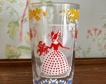 Könnte beinhalten: Klarer Glasbecher mit Vintage-Design. Das Glas zeigt eine Frau in einem roten Kleid mit weißem Polka-Dot-Rock, die einen Blumenstrauß hält. Gelbe Blumen- und blaue Blumenmuster verzieren die Ober- und Unterseite des Glases.