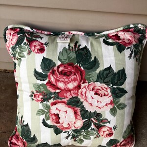 Puede incluir: Una almohada cuadrada con fondo blanco y rayas verdes y blancas. La almohada presenta un patr&oacute;n floral de rosas rosas y rojas con hojas verdes.