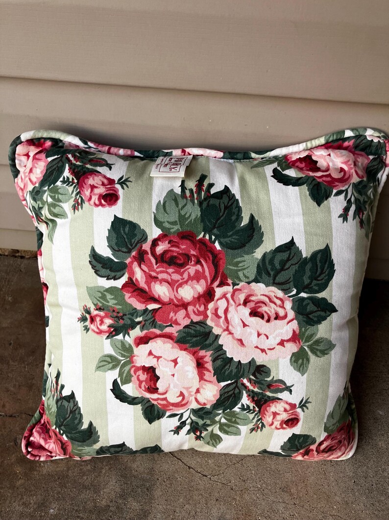Puede incluir: Una almohada cuadrada con fondo blanco y rayas verdes y blancas. La almohada presenta un patr&oacute;n floral de rosas rosas y rojas con hojas verdes.