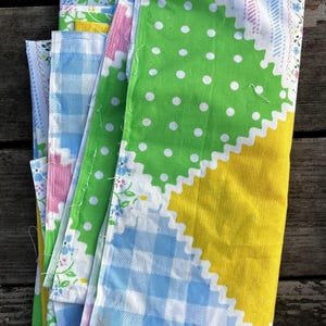 May include: A stack of colorful fabric pieces with various patterns. The fabrics feature green polka dots, blue gingham, yellow, pink, and floral designs. The edges have a scalloped trim. The fabrics are folded and stacked on a wooden surface.