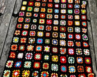 Könnte beinhalten: Eine rechteckige Häkeldecke mit einem Gitter aus bunten Granny Squares. Jedes Quadrat ist mit schwarzem Garn umrandet, was ein kontrastreiches Muster erzeugt. Die Quadrate enthalten verschiedene Farben wie Rot, Gelb, Blau, Grün und Weiß.