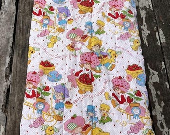 Könnte beinhalten: Ein rechteckiger Schlafsack aus weißem Stoff mit einem sich wiederholenden Muster aus Strawberry Shortcake-Figuren, gelben Wagen voller Erdbeeren und kleinen rosa Tieren. Der Text lautet Love is a Warm and Cozy Feeling.