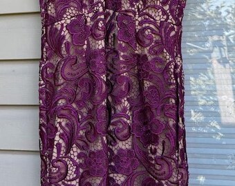 Könnte beinhalten: Ein ärmelloses, knielanges Kleid mit tiefviolettem floralem Spitzen-Overlay. Das Kleid hat einen V-Ausschnitt und ein nacktfarbenes Futter. Das Spitzendesign weist komplizierte Blumen- und Wirbelmuster auf.