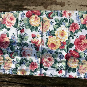 Puede incluir: Funda de almohada blanca con estampado floral. El diseño presenta grandes rosas en tonos rojos, amarillos y rosas, intercaladas con hojas verdes y pequeñas flores azules. La tela es probablemente de algodón o similar.