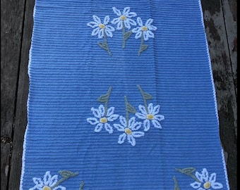 Könnte beinhalten: Ein rechteckiges blaues Handtuch mit gerippter Textur und weißem Rand. Das Handtuch ist mit gestickten Gänseblümchenmotiven verziert, die jeweils aus weißen Blütenblättern, gelben Mitten und grünen Stielen bestehen und in Gruppen angeordnet sind.