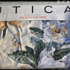 Puede incluir: Una sábana plana queen size empaquetada de Utica. La sábana presenta un estampado floral con grandes flores en tonos amarillos, blancos y azules, sobre un fondo crema. El embalaje dice "UTICA" y "ONE QUEEN FLAT SHEET".
