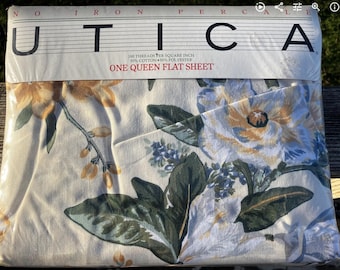 Könnte beinhalten: Ein verpacktes Queen-Size-Flachbettlaken von Utica. Das Laken zeigt ein Blumenmuster mit großen Blüten in Gelb-, Weiß- und Blautönen auf cremefarbenem Hintergrund. Die Verpackung trägt die Aufschrift "UTICA" und "ONE QUEEN FLAT SHEET".