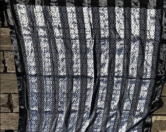 Könnte beinhalten: Ein quadratisches Tuch mit einem sich wiederholenden schwarz-silbernen Monogrammmuster in vertikalen Streifen. Das Tuch hat einen einfarbigen schwarzen Rand an allen vier Seiten und enthält das Wort Coach in kleiner Schrift oben links.