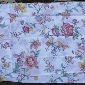 Pode incluir: Tecido branco com um padrão floral e de borboletas. O design apresenta flores rosa, amarelas e roxas com caules e folhas verdes. Borboletas amarelas e roxas estão espalhadas.