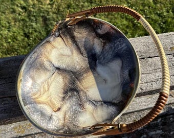 Könnte beinhalten: Eine runde, flache Schale mit einem wirbelnden, marmorierten Muster in Schwarz, Weiß und Metallic-Gold. Die Schale ist mit einem geschwungenen, bogenförmigen Griff ausgestattet, der mit abwechselnden Bändern aus hellbraunem und dunkelbraunem Flechtmaterial umwickelt ist.