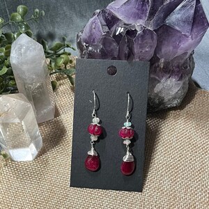 May include: A pair of dangle earrings with red gemstone drops and white opal accents. The earrings are made with silver wire and have a simple, elegant design.