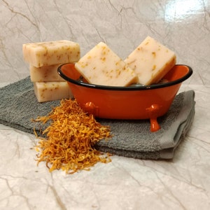 May include: Four bars of handmade soap with dried flowers in a small orange bathtub on a gray towel with dried orange flower petals scattered around the tub.