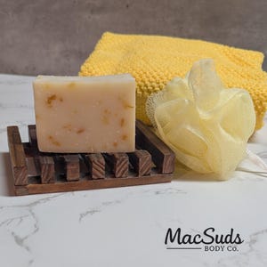 May include: A bar of soap with visible botanicals rests on a dark wooden soap dish. A yellow washcloth and a loofah are next to the soap. The soap has a light color with flecks of botanicals. The brand name "MacSuds BODY CO." is visible.