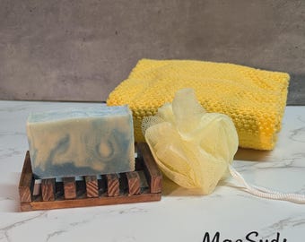 May include: A bar of blue and white marbled soap rests on a wooden soap dish, next to a yellow washcloth and a yellow loofah. The soap dish is made of dark brown wood. The text "MacSuds BODY CO." is at the bottom.