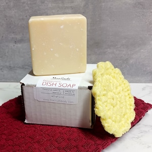 May include: A white box with a label that reads "MacSuds Peppermint DISH SOAP Coconut oil, castor oil, essential oil MADE IN MISSOURI NET WT. 6 OZ www.macsudsbody.com". A bar of white soap is sitting on top of the box. A yellow crocheted dish scrubber is sitting next to the box.