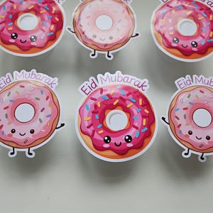 May include: Six donut-shaped stickers with the text "Eid Mubarak". Each sticker features a smiling donut with pink frosting and colorful sprinkles. The donuts have arms and legs.