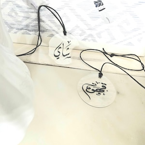 May include: Two clear acrylic pendants with black string necklaces. The pendants have Arabic calligraphy written on them. The top pendant says "Shakay" and the bottom pendant says "Habibi".