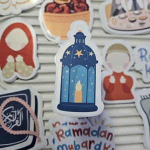 May include: A set of Ramadan themed stickers featuring a blue lantern with stars and a candle, a red and white prayer rug, a bowl of dates, a book with Arabic script, and a sticker that says "Ramadan Mubarak!"