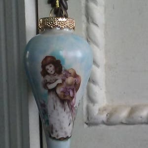 Könnte beinhalten: Hellblaue, tropfenförmige Weihnachtskugel mit goldfarbener Kappe und detaillierter Illustration einer Frau, die Blumen hält. Der Schmuck wird von einer goldenen Schnur und einem dunkelbraunen Dekorationselement gehalten. Die Website victoriandollslampsandmore.com ist unten aufgedruckt.