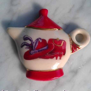 Puede incluir: Una tetera blanca pequeña con tapa y base rojas. La tetera está decorada con un sombrero rojo con plumas moradas y un zapato rojo.