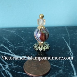 May include: A miniature glass perfume bottle with a gold-colored base and a swirl pattern in shades of pink, white, and black. The bottle has a clear glass stopper with a gold-colored top.