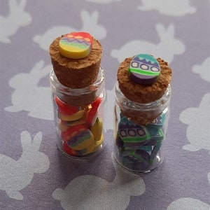 May include: Two small glass bottles with cork tops filled with colourful Easter egg shaped charms. The bottles are on a purple background with white bunny silhouettes.