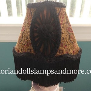 May include: A lampshade with a black fringe and a gold and red patterned fabric. The fabric has a paisley design with a black velvet center.