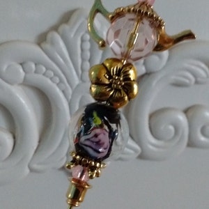 May include: A gold teapot charm with pink and clear glass beads, a gold flower bead, and a clear glass bead with a pink and black flower design. The charm is attached to a gold pin.