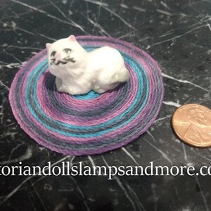 Può includere: Una piccola statuina di gatto in ceramica bianca seduta su un tappeto rotondo, colorato e multistrato. Il tappeto ha sfumature di viola, blu e rosa.