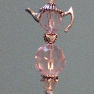 May include: A silver and pink teapot charm with a heart and clear beads. The charm is attached to a silver pin.