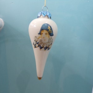 Puede incluir: Adorno navideño blanco en forma de lágrima con un diseño de bufón azul y dorado. Tiene una tapa dorada y una parte superior decorativa azul. El fondo es una pared azul claro.