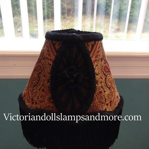 May include: A gold and red patterned lampshade with black fringe and trim. The lampshade has a black fabric bow in the center.