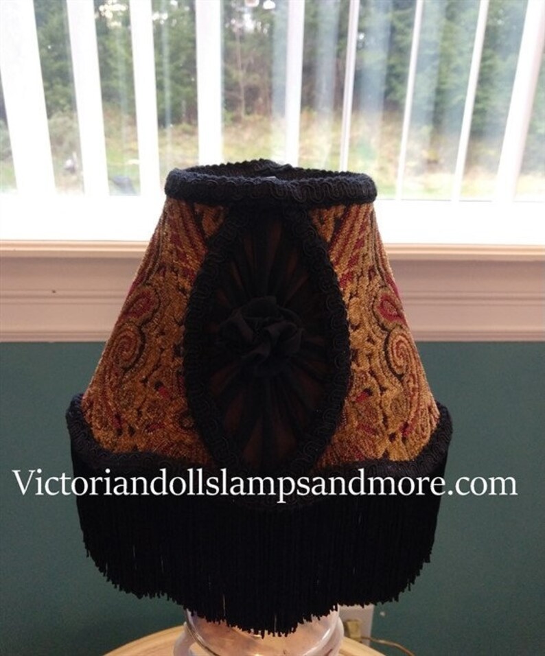 May include: A gold and red patterned lampshade with black fringe and trim. The lampshade has a black fabric bow in the center.