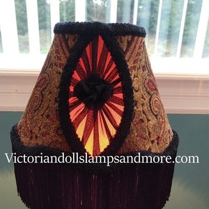 May include: A lampshade with a black fringe and a gold and red patterned fabric. The shade has a black ribbon with a red and black fan-shaped design in the center. The text "victoriandollslampsandmore.com" is visible at the bottom of the image.