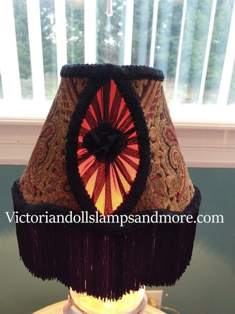 May include: A lampshade with a black fringe and a gold and red patterned fabric. The shade has a black ribbon with a red and black fan-shaped design in the center. The text "victoriandollslampsandmore.com" is visible at the bottom of the image.