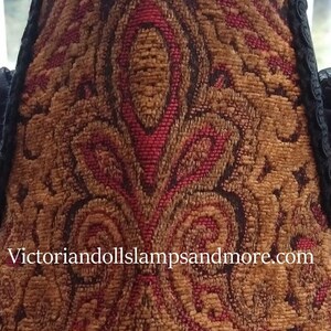 May include: A close-up of a lampshade with a red and gold paisley pattern. The fabric is textured and has a black trim. The website address "victoriandollslampsandmore.com" is printed on the bottom of the lampshade.