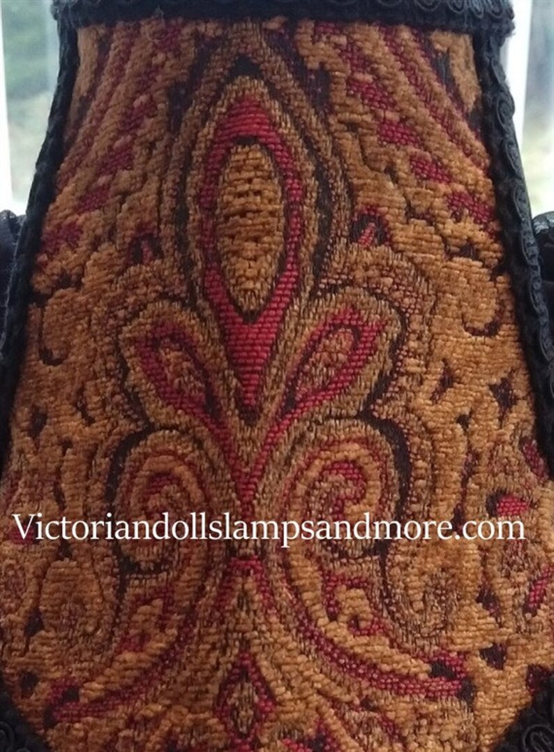 May include: A close-up of a lampshade with a red and gold paisley pattern. The fabric is textured and has a black trim. The website address "victoriandollslampsandmore.com" is printed on the bottom of the lampshade.