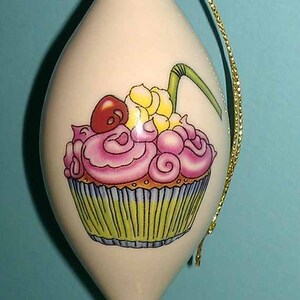 Może przedstawiać: Biała ceramiczna ozdoba choinkowa w kształcie łzy ze złotym wierzchołkiem i ręcznie malowanym wzorem ciasta. Ciasto ma różowy lukier, czerwoną wiśnię i żółte posypki.